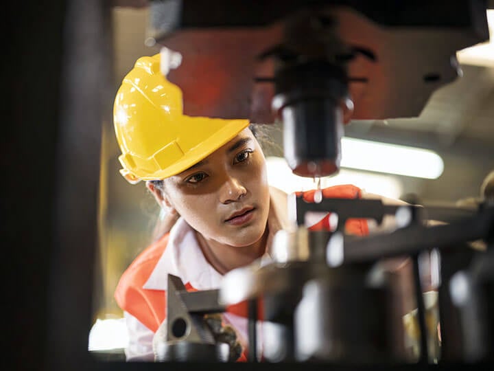 Female with yellow hardhat and orange caution vest using a drill press