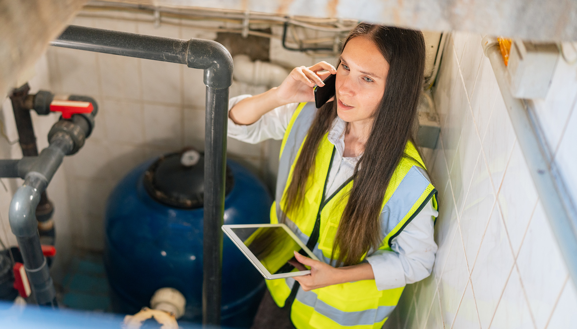 female worker in PPE on site, on phone, with tablet, attending to water pipe system