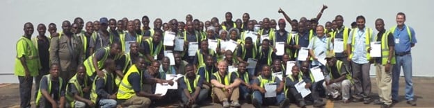 A happy group of multiple delegates in sunshine, wearing hi-vis vests, many holding certificates.